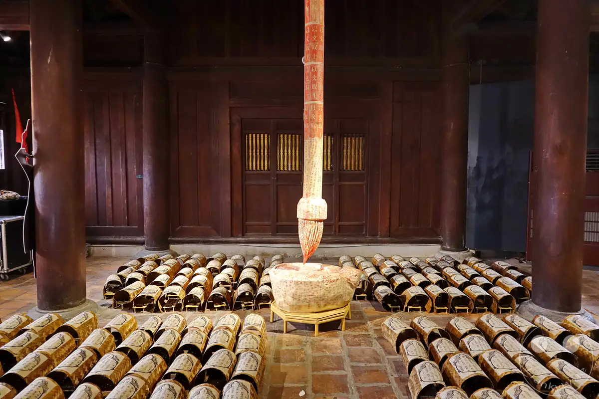 Im Inneren eines traditionellen Gebäudes in Hanoi, Vietnam, werden Dutzende von Artefakten aus geflochtenem Material, die mit Schriftzeichen verziert sind, in Reihen auf dem Boden ausgestellt. Ein zentrales Kunstwerk, das wie ein riesiger Pinsel mit Schriftzeichen verziert ist, dominiert die Szene vor einer holzvertäfelten Wand. Die Atmosphäre ist ruhig und ehrfürchtig, was auf eine kulturelle oder historische Ausstellung im Tempel der Literatur hindeutet.