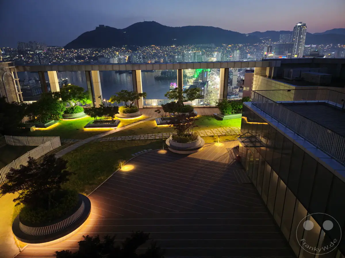 Nachts bietet sich ein atemberaubender Panoramablick von der Dachterrasse des LOTTE Department Store in Busan, Südkorea. Die beleuchtete Stadtlandschaft erstreckt sich bis zu den sanften Hügeln im Hintergrund, während die künstliche Beleuchtung des Gartens und der Architektur für eine stimmungsvolle Atmosphäre sorgt. Das Bild fängt die friedliche Stille der Nacht in einer pulsierenden Metropole ein.
