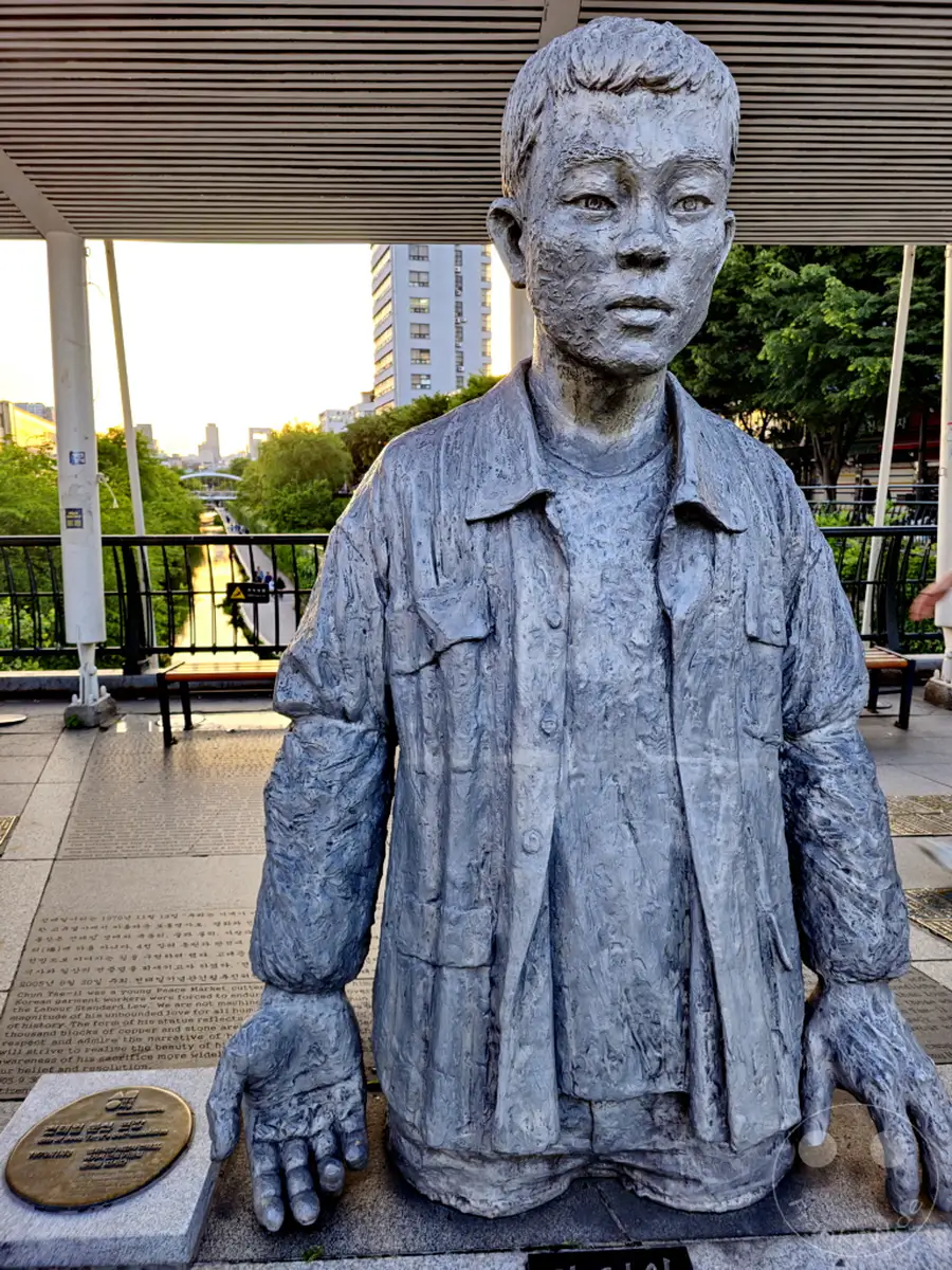 Die Statue von Arbeitern, die die Rechte der Arbeiter repräsentieren, steht am Ufer des Han-Flusses in Seoul, Südkorea, nahe der Beodeuldari Bridge. Ein junger Mann, dargestellt als Statue, blickt nachdenklich in die Ferne, während im Hintergrund die Skyline der Stadt und grüne Bäume zu sehen sind. Die Skulptur strahlt eine ernste und nachdenkliche Atmosphäre aus, die die Bedeutung von Arbeiterrechten unterstreicht.