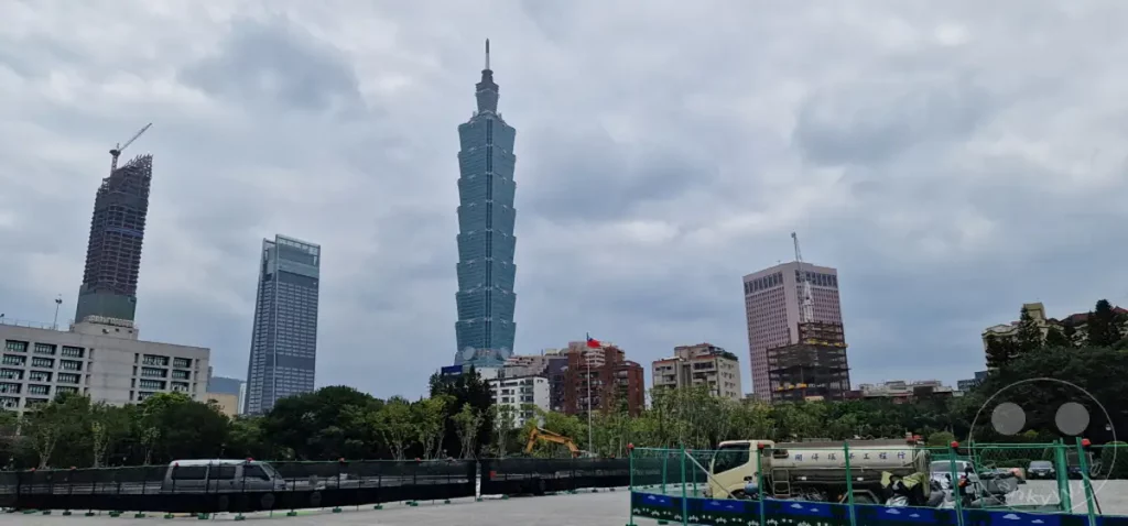 In Taipeh, Taiwan, ragen mehrere beeindruckende Wolkenkratzer, darunter der ikonische Taipei 101, unter einem bewölkten Himmel empor. Eine Baustelle im Vordergrund mit einem LKW und Bauzäunen deutet auf stetige Stadtentwicklung hin. Die Atmosphäre wirkt leicht melancholisch, aber auch dynamisch durch die sichtbare Konstruktion der urbanen Zukunft.