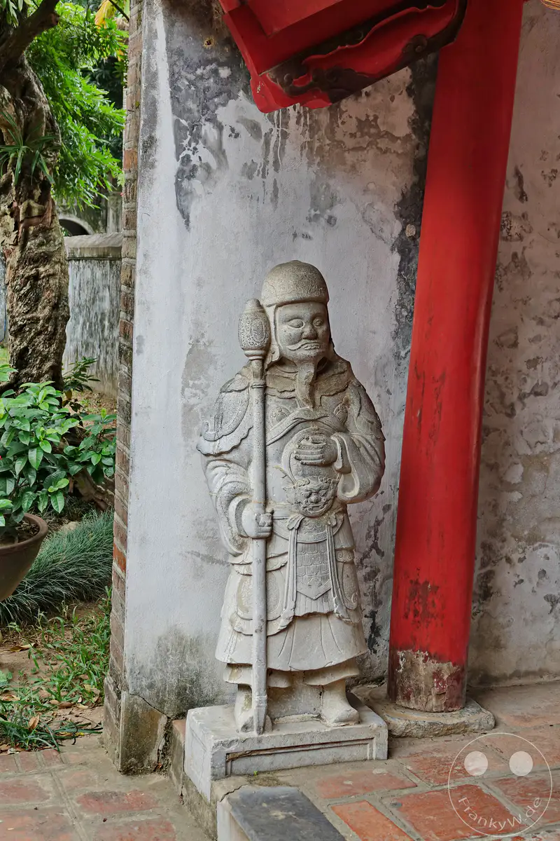 Eine steinerne Statue eines Wächters steht vor einer verputzten Wand im Literaturtempel in Hanoi, Vietnam. Eine leuchtend rote Säule und üppige grüne Vegetation umgeben die Skulptur und schaffen eine Atmosphäre der Ruhe und des historischen Erbes. Der detailreiche Stein und die architektonischen Elemente deuten auf die tief verwurzelten Traditionen und die reiche Kultur des Ortes hin.