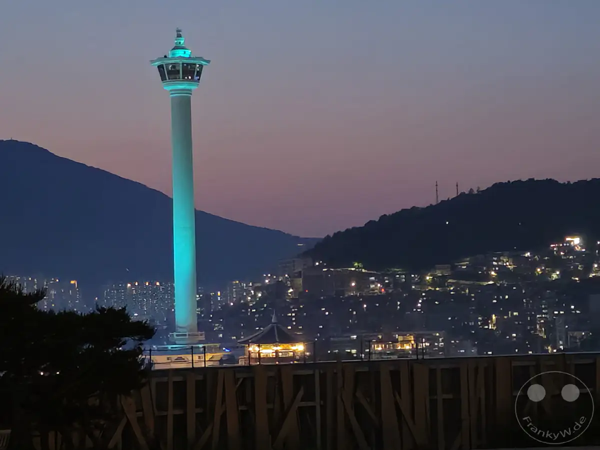 Der Busan Tower leuchtet in einem türkisen Farbton vor dem lila-orangenen Abendhimmel. Lichter der Stadt erhellen die Hügel und den Hintergrund, während am unteren Bildrand eine Holzkonstruktion zu sehen ist. Die Stimmung ist ruhig und friedlich mit einem Blick auf die nächtliche Skyline von Busan.