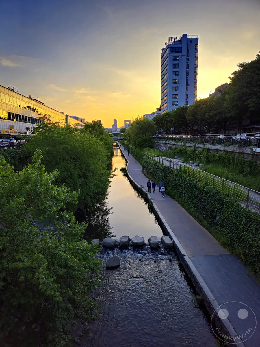 Der Cheonggyecheon Stream fließt durch Seoul in Südkorea, gesäumt von grünen Ufern und einem Fußweg. Eine Gruppe von Menschen spaziert entlang des Ufers, während die Abendsonne den Himmel in warme Farben taucht und die moderne Architektur der Stadt beleuchtet. Kleine Steine überqueren den Fluss und schaffen eine malerische Szenerie bei Sonnenuntergang.