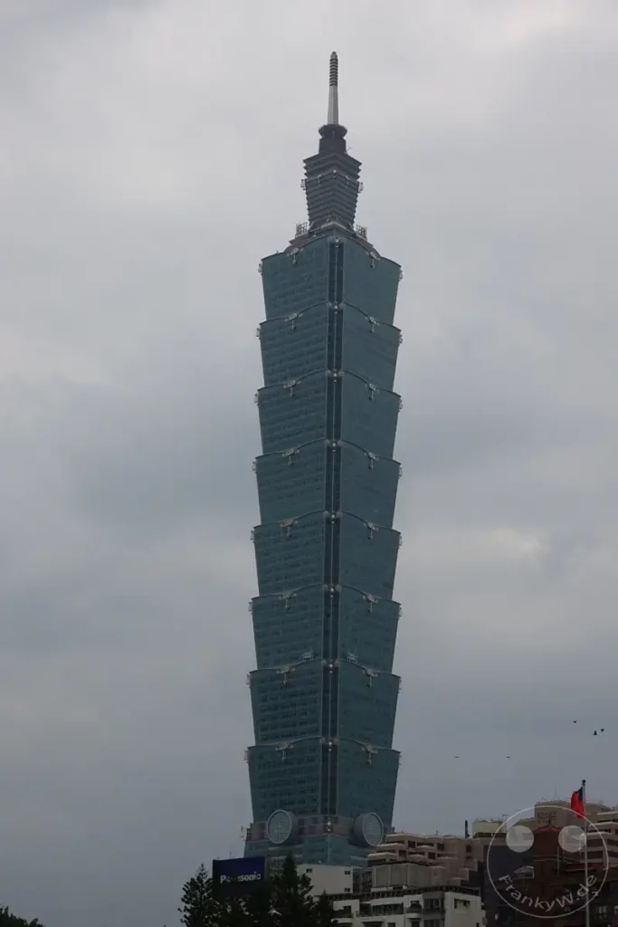 Das Wahrzeichen Taipehs, der Taipei 101, ragt imposant in den grauen Himmel. Die charakteristische Architektur des Wolkenkratzers wird durch die dramatische Wolkendecke betont. Unterhalb des Turms sind weitere Gebäude und Bäume zu erkennen, die die städtische Kulisse vervollständigen.