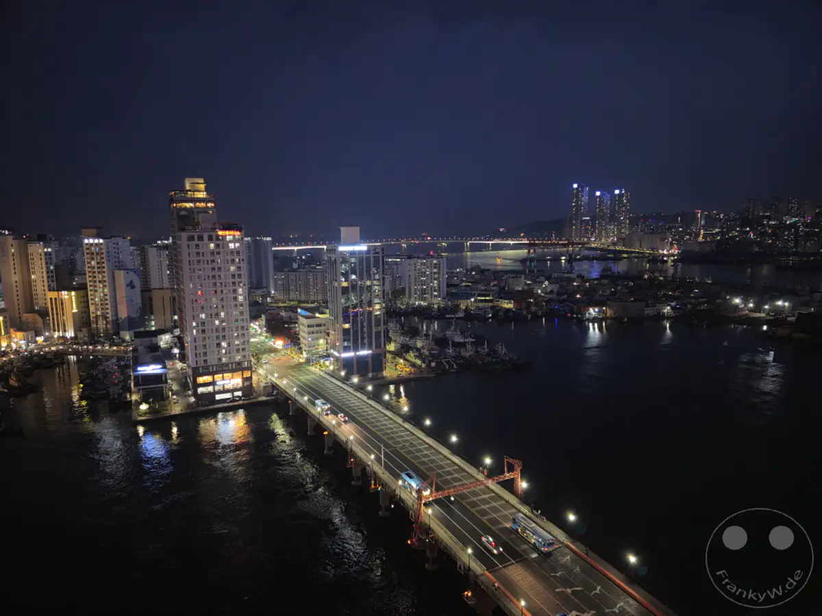 Die beleuchtete Skyline von Busan erstreckt sich über das dunkle Wasser, während Autos über eine Brücke fahren und die Lichter der Stadt reflektieren. Ein Panoramablick von einer erhöhten Position enthüllt die nächtliche Geschäftigkeit und architektonische Pracht. Die Szene strahlt eine ruhige, aber lebendige Atmosphäre aus, typisch für eine moderne Metropole nach Sonnenuntergang.