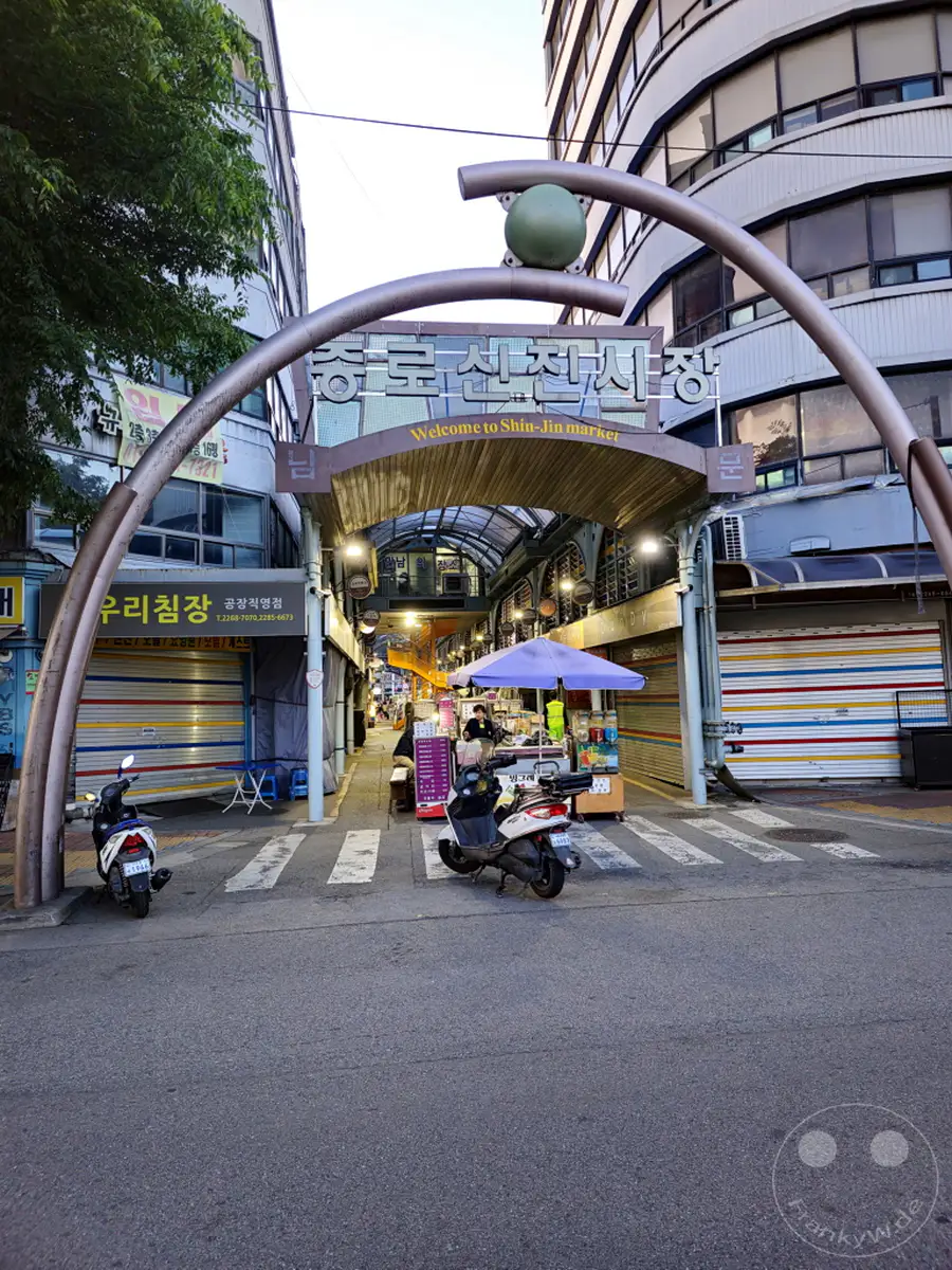 Der Eingang zum Jongno Shinjin Market in Seoul ist mit einem geschwungenen Metallbogen und einer grünen Kugel als Dekoration versehen. Roller sind auf der Straße abgestellt, während Geschäfte entlang der überdachten Marktstraße ihre Rollläden heruntergelassen haben. Die Szene vermittelt eine ruhige Atmosphäre kurz vor der Öffnung oder nach Geschäftsschluss.