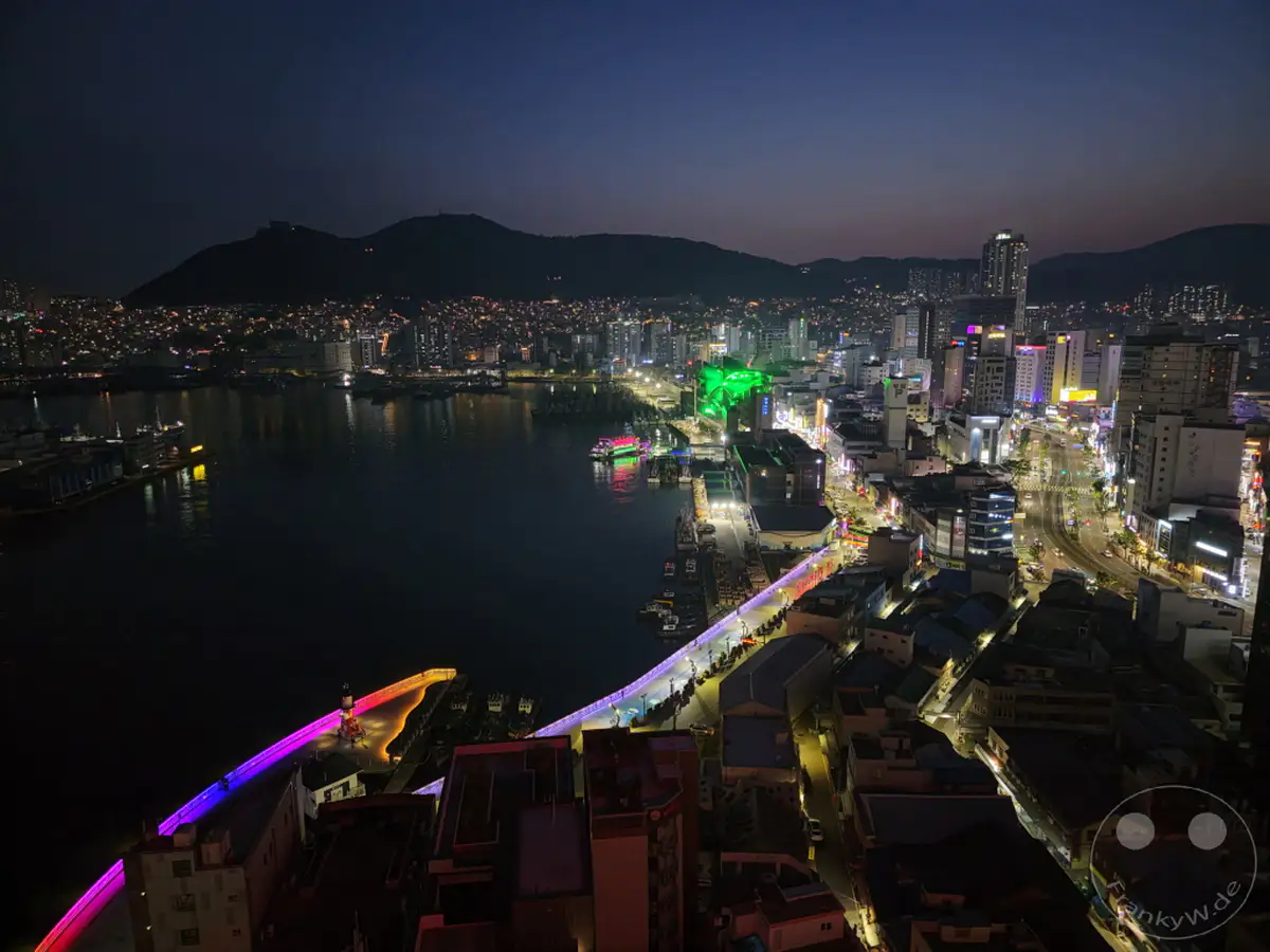 Die nächtliche Stadtlandschaft von Busan, Südkorea, erstreckt sich unter einem dunklen Himmel, beleuchtet von zahlreichen Lichtern. Der belebte Hafenbereich mit Schiffen und die bunte Beleuchtung einer Uferpromenade dominieren die Szene. Hochhäuser und Hügel im Hintergrund komplettieren den eindrucksvollen Ausblick bei Nacht.