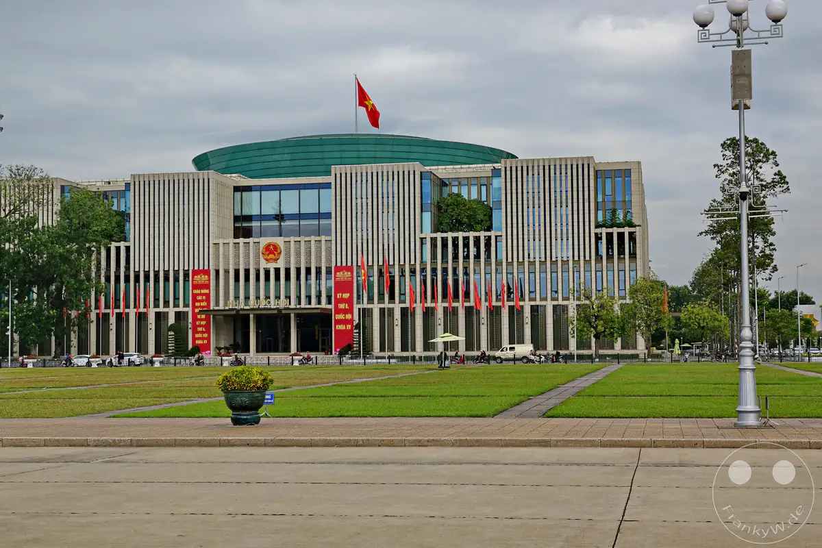 Das National Assembly Building in Hanoi, Vietnam, ragt mit seiner modernen Architektur und der grünen, kuppelförmigen Dachkonstruktion imposant in den bewölkten Himmel. Rote Fahnen und das vietnamesische Nationalemblem schmücken die Fassade, während davor gepflegte Rasenflächen und eine belebte Straße mit Fahrzeugen und Fußgängern zu sehen sind. Die Szene strahlt eine offizielle und zugleich geschäftige Atmosphäre aus, charakteristisch für das Zentrum politischer Macht.