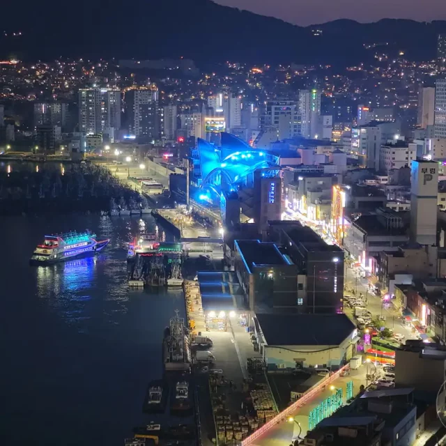Die Skyline von Busan erstrahlt bei Nacht mit unzähligen Lichtern, die eine lebendige Atmosphäre schaffen. Ein beleuchtetes Schiff gleitet durch den Hafen, während die Stadt und die umliegenden Berge im Dunkel der Nacht verschwinden. Die Szenerie bietet einen faszinierenden Blick auf das städtische Leben und die maritime Aktivität in Südkorea.