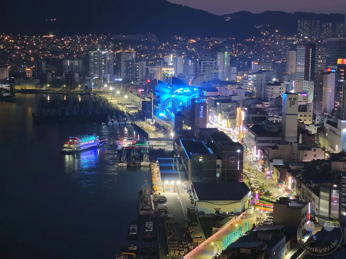 Die Skyline von Busan erstrahlt bei Nacht mit unzähligen Lichtern, die eine lebendige Atmosphäre schaffen. Ein beleuchtetes Schiff gleitet durch den Hafen, während die Stadt und die umliegenden Berge im Dunkel der Nacht verschwinden. Die Szenerie bietet einen faszinierenden Blick auf das städtische Leben und die maritime Aktivität in Südkorea.