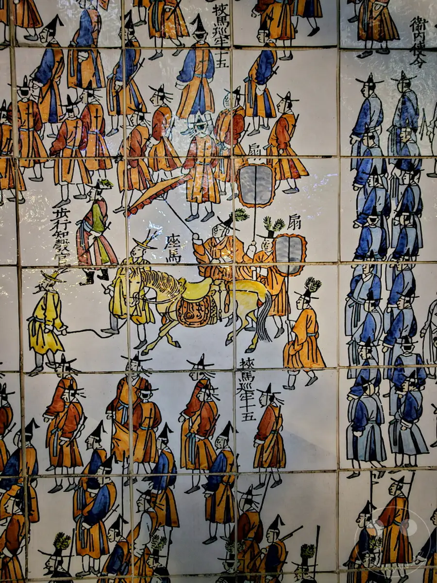 Detailreiche Keramikmalerei aus Südkorea, die eine historische Prozession darstellt. Die aufwendig gestalteten Figuren und Szenen am Ufer des Cheonggyecheon Streams in Seoul erwecken die Vergangenheit zum Leben. Die lebendigen Farben und die feinen Linienführung vermitteln einen Eindruck von festlicher oder zeremonieller Stimmung.