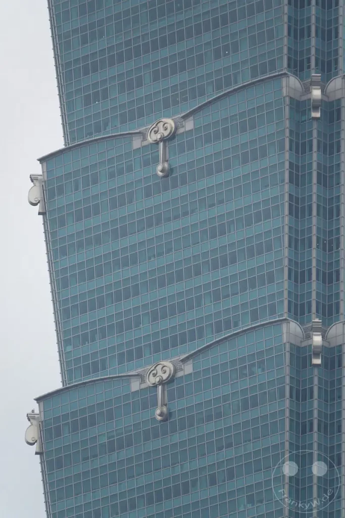 Die elegante Fassade des Taipei 101 Wolkenkratzers in Taipeh, Taiwan, präsentiert sich in einem kühlen Blauton. Die symmetrisch angeordneten architektonischen Elemente betonen die vertikale Struktur des Gebäudes. Eine gedämpfte Atmosphäre wird durch den bewölkten Himmel und die ruhige Darstellung der modernen Architektur erzeugt.