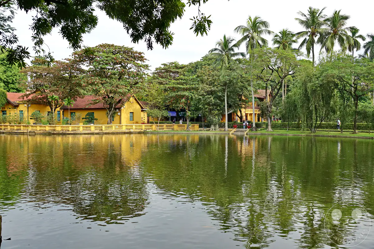 In Hanoi, Vietnam, erstreckt sich ein ruhiger Karpfenteich vor der historischen Kulisse des Hauses von Ho Chi Minh. Tropische Bäume spiegeln sich im sanft bewegten Wasser und schaffen eine friedliche Atmosphäre. Die Szenerie lädt zum Verweilen und Entspannen inmitten der Natur und Geschichte ein.