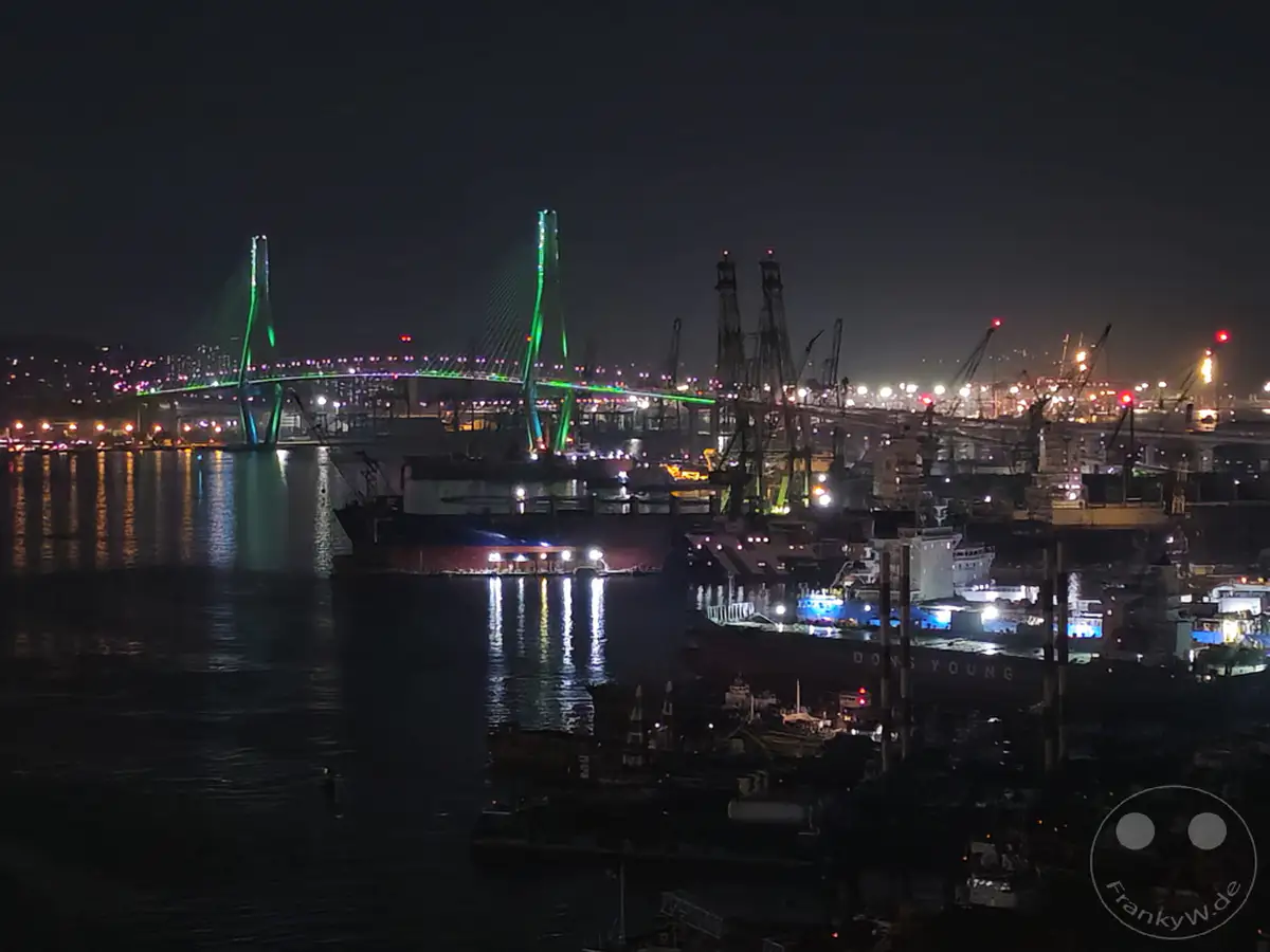 Die beleuchtete Brücke von Busan erstreckt sich über das nächtliche Wasser und reflektiert ihr grünes Licht. Am Ufer ist ein geschäftiger Hafen mit Kränen und Schiffen zu sehen, die eine belebte Industrielandschaft bilden. Die Szene vermittelt eine pulsierende und zugleich ruhige Atmosphäre der Stadt bei Nacht.
