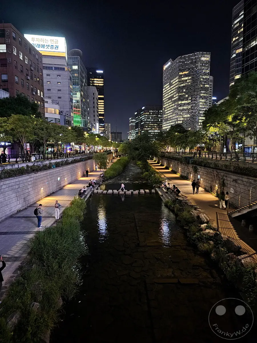 Der beleuchtete Cheonggyecheon Stream in Seoul, Südkorea, wird nachts von den umliegenden Wolkenkratzern erhellt. Spaziergänger genießen die ruhige Atmosphäre entlang des Wasserlaufs und der Steintreppen, die den Fluss überqueren. Die Szene bietet einen malerischen Kontrast zwischen urbaner Beleuchtung und natürlicher Wasserlandschaft.