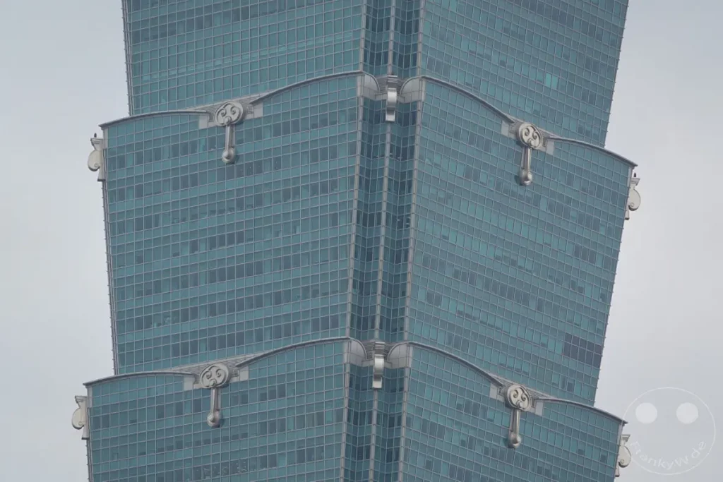 Die markante Fassade des Taipei 101 Wolkenkratzers in Taipeh, Taiwan, präsentiert sich mit seinen charakteristischen, sich nach oben verjüngenden Segmenten und den symmetrisch angeordneten architektonischen Verzierungen. Die strukturellen Elemente und die bläulich getönten Glasflächen vermitteln einen Eindruck von fortschrittlicher Ingenieurskunst und urbaner Eleganz. Der graue Himmel und die Fokussierung auf die Architektur erzeugen eine kühle und imposante Atmosphäre.