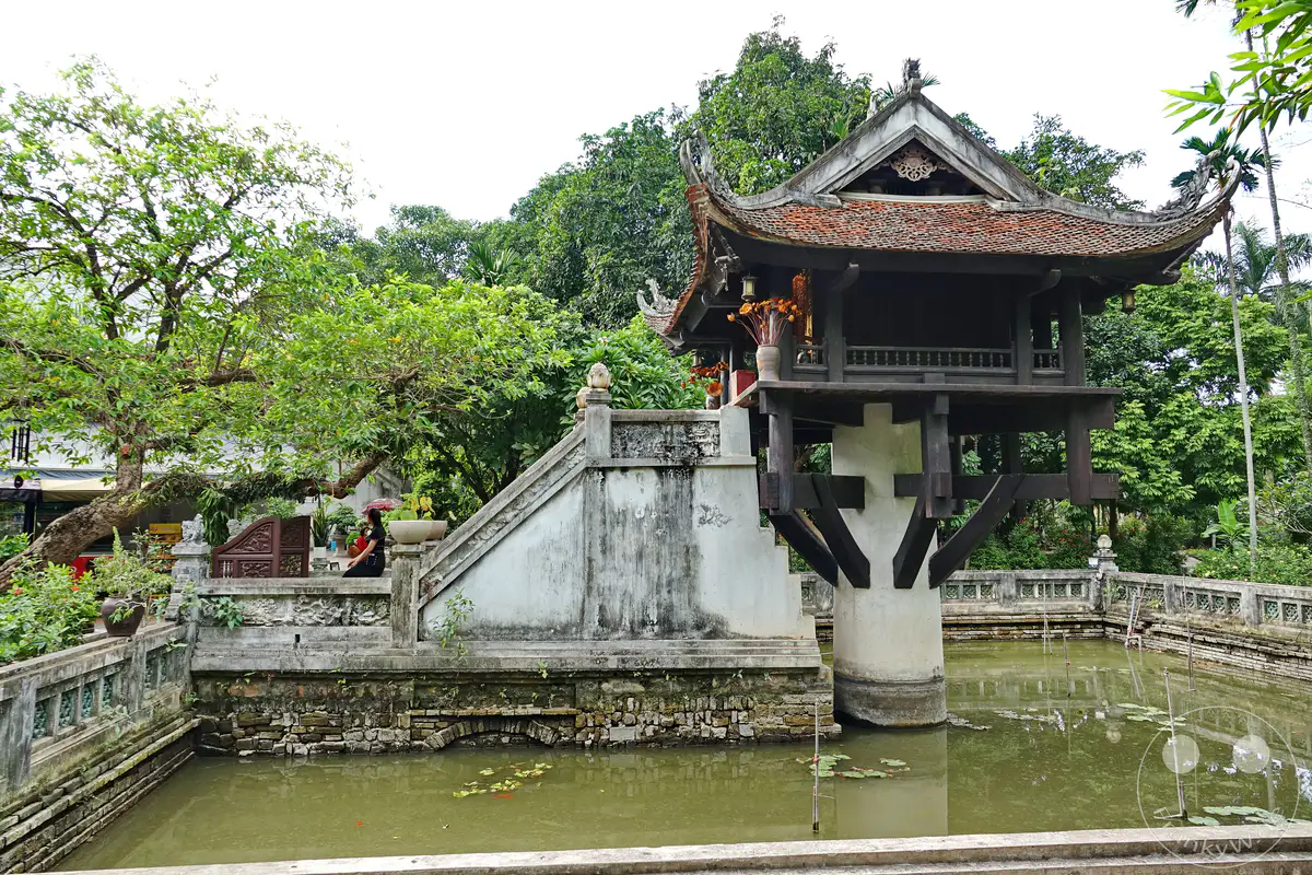Ein steinerner Teich mit Seerosen und zwei Koi-Karpfen bildet die Basis für die berühmte Ein-Säulen-Pagode in Hanoi, Vietnam. Die Pagode mit ihrem traditionellen vietnamesischen Dach und den kunstvollen Verzierungen steht auf einem einzelnen dicken Baumstamm im Wasser. Umgeben von üppiger Vegetation und Bäumen strahlt die Szenerie eine friedliche und historische Atmosphäre aus.