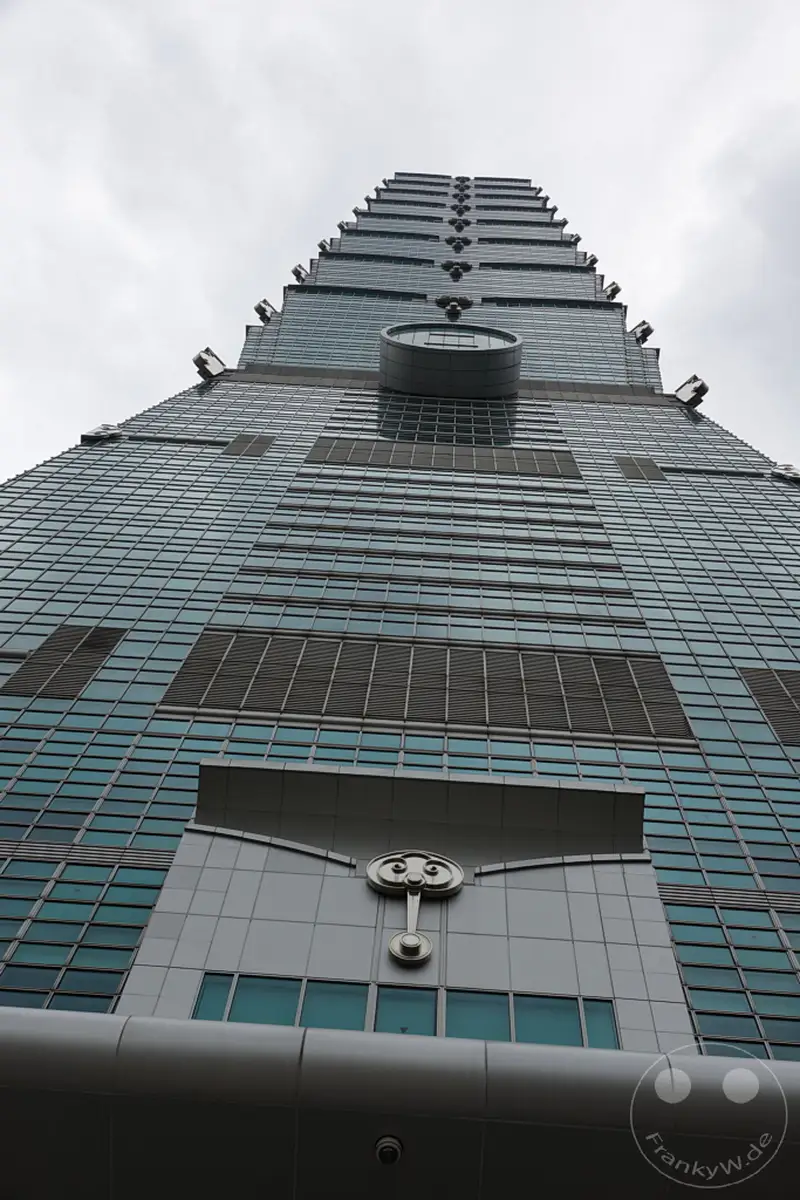Der Blick nach oben auf den imposanten Taipei 101 in Taiwan fängt die architektonische Meisterleistung dieser modernen Struktur ein. Die gläserne Fassade reflektiert den grauen Himmel und verleiht dem Gebäude eine majestätische Präsenz inmitten der urbanen Landschaft von Taipeh. Die vertikale Ausrichtung und die symmetrische Form unterstreichen die geometrische Präzision und die beeindruckende Höhe des Wolkenkratzers.