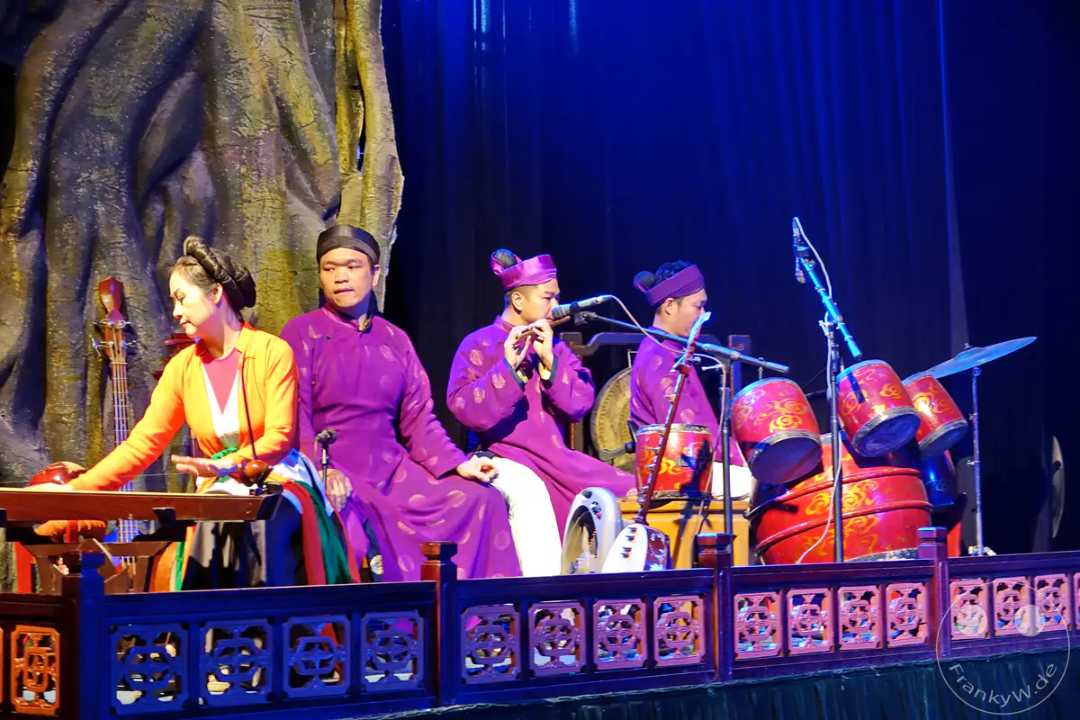Eine Gruppe Musiker spielt traditionelle vietnamesische Musik auf einem Holzpodest, das mit Schnitzereien verziert ist, während das Bühnenbild einen dramatischen, baumartigen Hintergrund und tiefblaue Vorhänge zeigt. Die Darsteller, gekleidet in prächtige lila Roben, spielen verschiedene Instrumente, darunter eine Flöte und Trommeln, was eine lebendige und kulturelle Atmosphäre schafft. Dieses kulturelle Spektakel findet wahrscheinlich in Hanoi statt und präsentiert das reiche Erbe der vietnamesischen Wasserpuppenspieltradition.
