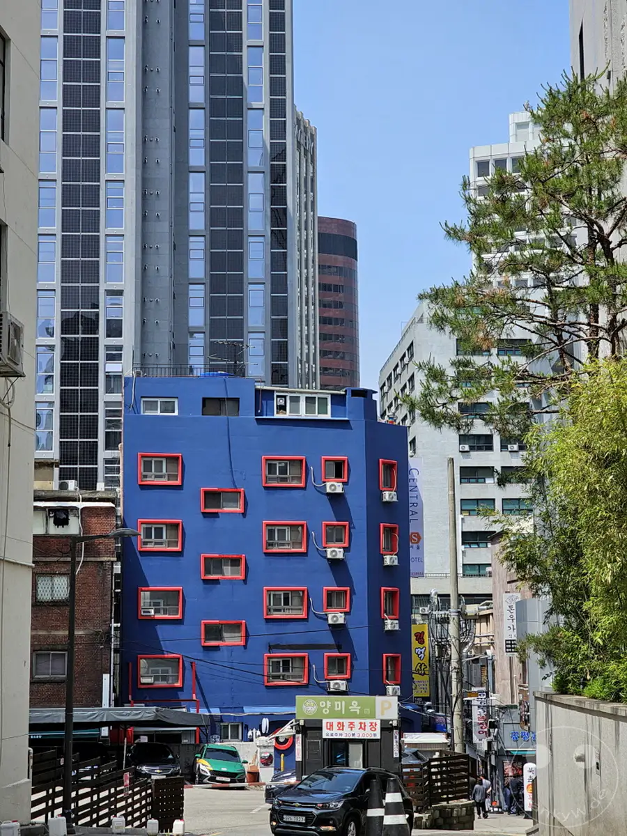 Ein leuchtend blaues Gebäude mit roten Fensterrahmen sticht in der urbanen Landschaft von Seoul, Südkorea, hervor und kontrastiert mit den umliegenden modernen Hochhäusern. Klimaanlagen sind an der Fassade des auffälligen Gebäudes angebracht, was auf eine Mischung aus künstlerischem Design und Funktionalität hinweist. Die Szene vermittelt eine lebendige und dynamische Atmosphäre inmitten des geschäftigen Stadtlebens.
