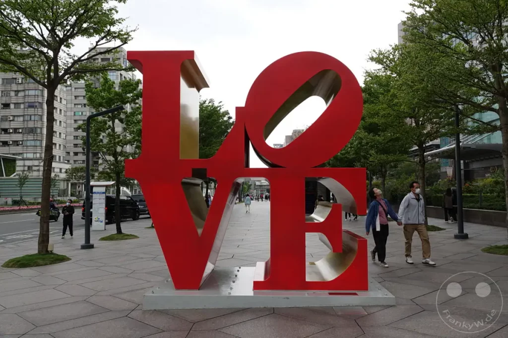 Eine ikonische rote LOVE-Skulptur steht im Vordergrund auf einem gepflasterten Platz in Taipeh, Taiwan. Im Hintergrund ist die Skyline der Stadt mit modernen Gebäuden und Bäumen zu sehen, während einige Menschen im Vordergrund spazieren gehen. Die Szene vermittelt ein Gefühl urbaner Lebendigkeit vor dem Hintergrund einer bekannten Sehenswürdigkeit.