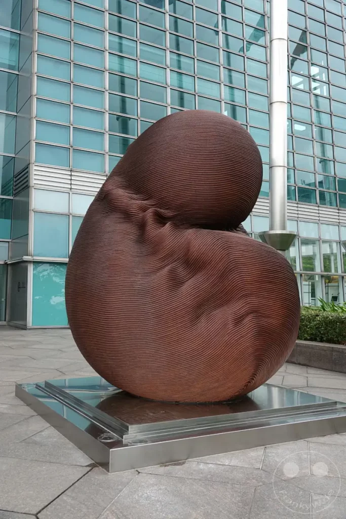 Eine moderne Außenskulptur aus braunem, geripptem Material steht auf einem polierten Metallsockel vor der gläsernen Fassade des Taipei 101 Gebäudes in Taipeh, Taiwan. Die organisch geformte Skulptur bildet einen interessanten Kontrast zur strengen Geometrie des Hochhauses. Die Szene vermittelt einen Eindruck von städtischer Kunst und Architektur.