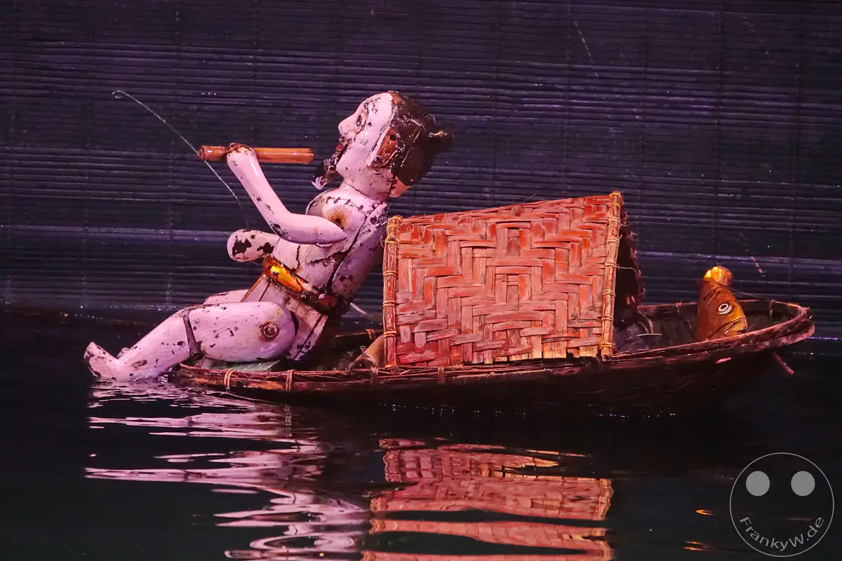 Eine kunstvoll gefertigte Marionette, die eine Fischerfigur darstellt, sitzt in einem traditionellen Boot auf dunklem Wasser. Die detailreiche Rattan-Konstruktion des Bootes und die antike Aufmachung der Puppe erzeugen eine atmosphärische Szene, die an vietnamesische Wassermarionettenspiele erinnert. Dieses Bild fängt die Essenz eines kulturellen Erbes im Herzen von Hanoi ein.