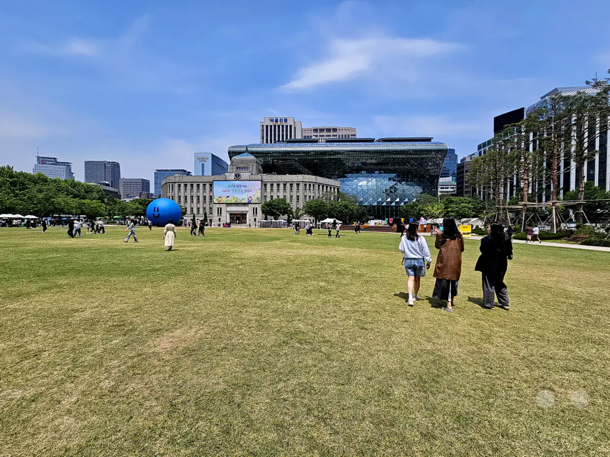 Menschen genießen einen sonnigen Tag auf dem weitläufigen Seoul Plaza, einem zentralen öffentlichen Raum in der südkoreanischen Hauptstadt Seoul. Umgeben von moderner Architektur und historischen Gebäuden bietet der Platz eine grüne Oase für Erholung und Veranstaltungen. Die lockere Atmosphäre lädt zum Verweilen und Beobachten des städtischen Treibens ein.