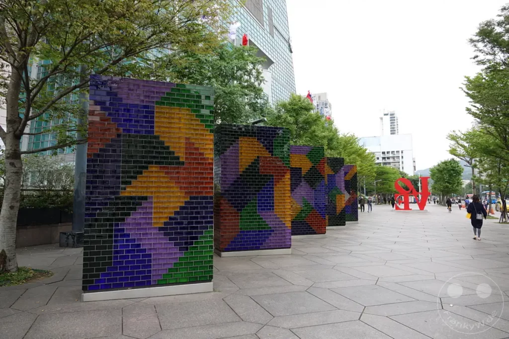 Eine Promenade in Taipeh, Taiwan, gesäumt von bunten, geometrischen Skulpturen, die einen starken visuellen Kontrast zur modernen Architektur bilden. Die breite, gepflasterte Fläche lädt zum Flanieren ein, während im Hintergrund die Skyline mit dem markanten Taipei 101 in Erscheinung tritt. Die Szene vermittelt eine Atmosphäre urbaner Kunst und öffentlicher Raumgestaltung an einem belebten Ort.