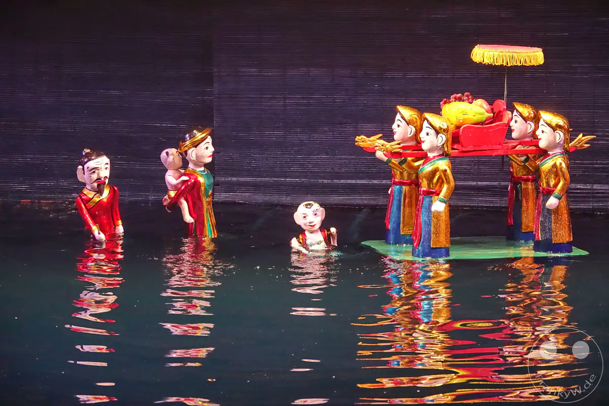 Wassermarionetten-Theater in Hanoi, Vietnam, wo traditionelle Figuren auf einer Wasserbühne interagieren. Die leuchtenden Farben der Kostüme und die Spiegelungen im Wasser erzeugen eine lebendige und magische Atmosphäre. Dieses kulturelle Erbe wird durch kunstvolle Aufführungen lebendig gehalten.