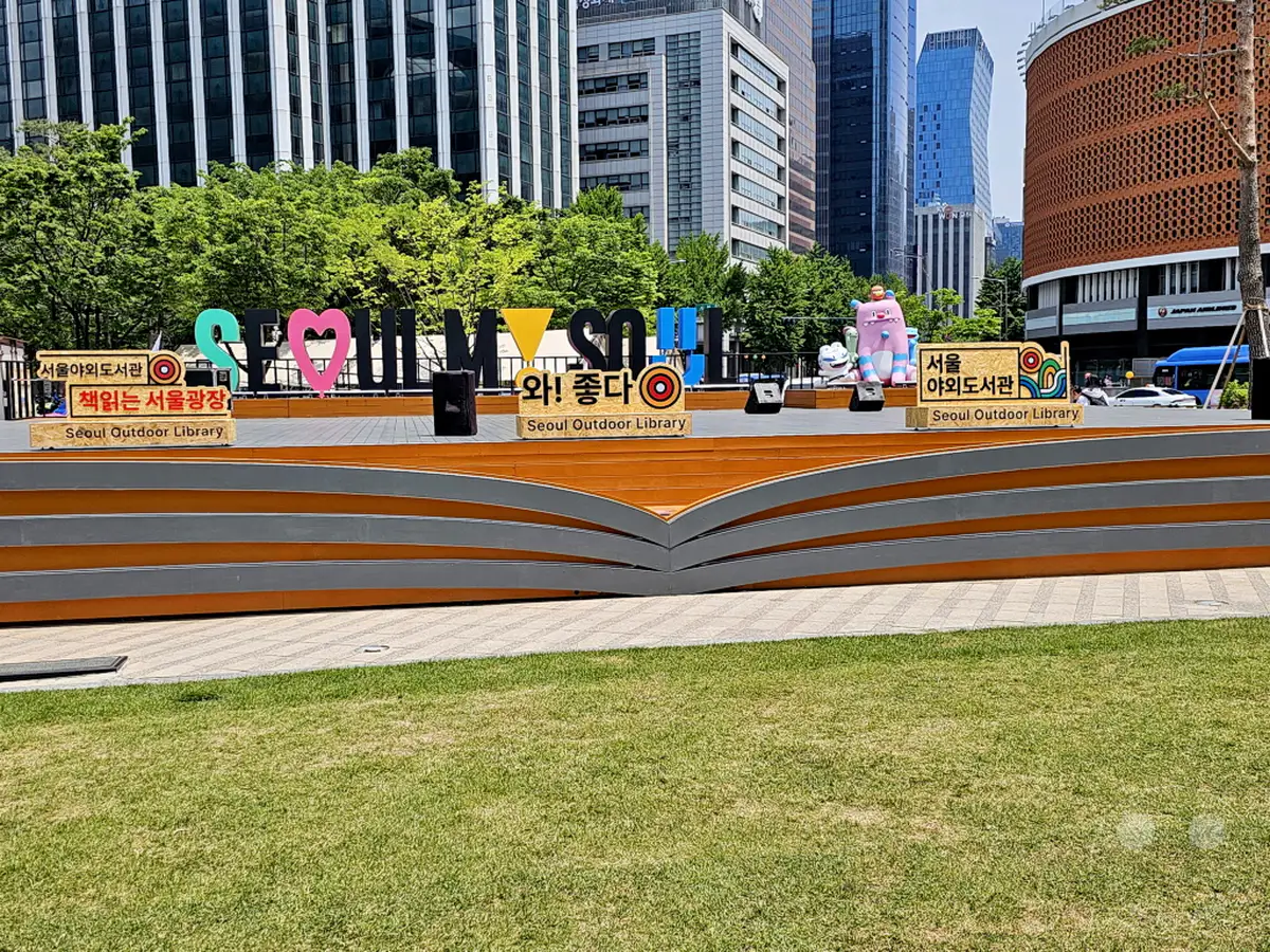 Ein heller, sonniger Tag unterstreicht die lebendige Atmosphäre in Seoul, Südkorea. Die "Seoul Outdoor Library" auf dem Seoul Plaza präsentiert sich als kreativer Treffpunkt, umgeben von moderner Stadtarchitektur und üppigem Grün. Die bunte Installation mit den Buchstaben und Figuren lädt zum Verweilen und Entdecken ein.