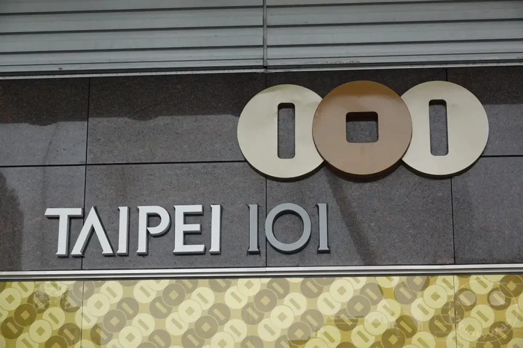 Das imposante Schild "TAIPEI 101" ist auf einem Granit-Fassadenbereich angebracht. Über dem Schriftzug sind drei stilisierten Kreiselemente zu sehen, die das Logo des Gebäudes bilden. Darunter zieren Muster aus weiteren Kreisen und Quadraten die Fassade.