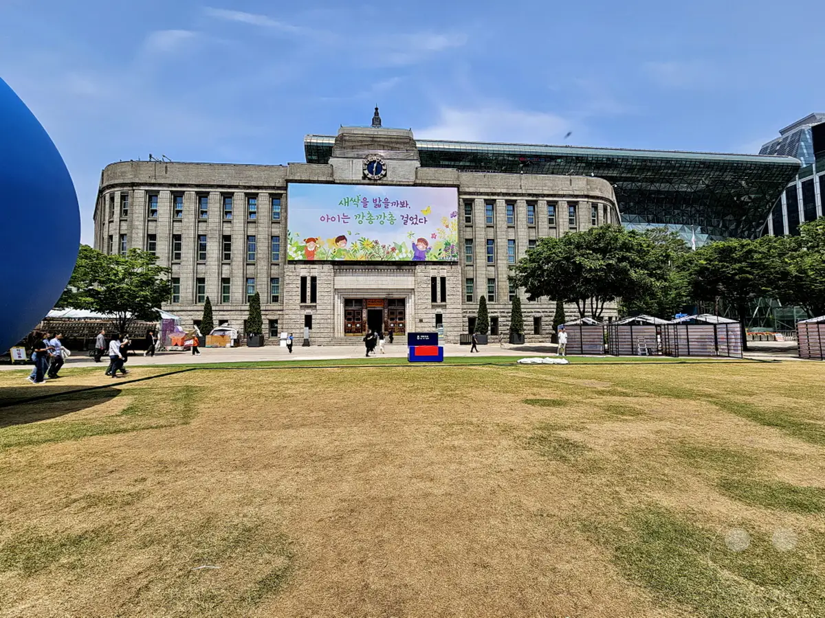 Südkorea, Seoul: Das Seoul Plaza ist von einer Mischung aus historischer und moderner Architektur geprägt, wobei ein großes Gebäude mit einem imposanten Bildschirm im Vordergrund steht. Auf dem Platz finden verschiedene Aktivitäten und Veranstaltungen statt, die von einer lebendigen Atmosphäre zeugen. Die Szene wird von strahlendem Sonnenschein und einer geschäftigen Kulisse belebt.