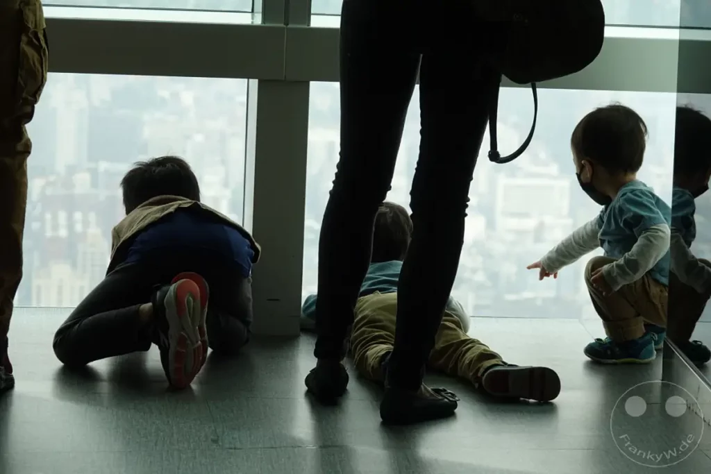 Eine Familie blickt von einem hohen Gebäude in Taipeh, Taiwan, aus der Vogelperspektive auf die Stadt. Kinder sitzen oder liegen auf dem Boden und schauen fasziniert aus dem Fenster. Die gedämpfte Beleuchtung und der Blick auf die weitläufige Metropole erzeugen eine ruhige und kontemplative Atmosphäre.