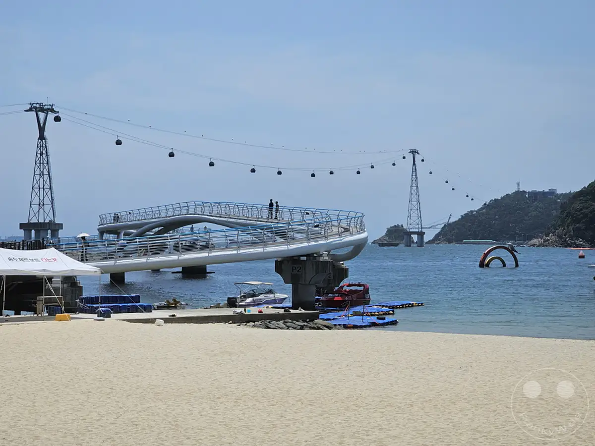 Eine sonnige Aufnahme von Südkorea, genauer gesagt von Busan, am Songdo Beach. Eine moderne Fußgängerbrücke erstreckt sich über das ruhige Meer, während die Seilbahn, die Songdo Cable Car, in der Ferne über die Bucht schwebt. Die friedliche Szenerie mit dem Sandstrand und dem tiefblauen Wasser lädt zum Entspannen ein.