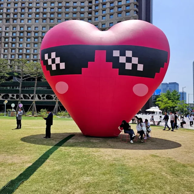 Ein riesiger, roter Herz-Ballon mit Pixel-Sonnenbrille steht auf einer grünen Rasenfläche in Seoul Plaza, Südkorea. Mehrere Personen sind in der Nähe des Ballons zu sehen, einige sitzen auf Stühlen und entspannen sich, während andere im Hintergrund spazieren gehen. Die Szene strahlt eine entspannte und festliche Atmosphäre an einem sonnigen Tag aus.