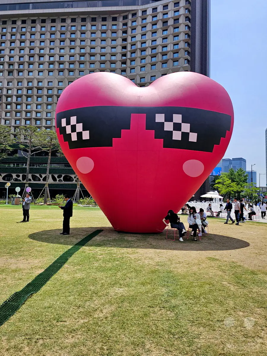 Ein riesiger, roter Herz-Ballon mit Pixel-Sonnenbrille steht auf einer grünen Rasenfläche in Seoul Plaza, Südkorea. Mehrere Personen sind in der Nähe des Ballons zu sehen, einige sitzen auf Stühlen und entspannen sich, während andere im Hintergrund spazieren gehen. Die Szene strahlt eine entspannte und festliche Atmosphäre an einem sonnigen Tag aus.