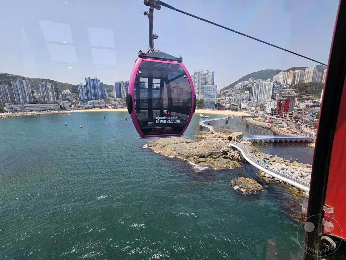Die Fahrt mit der Songdo Cable Car in Busan, Südkorea, bietet einen atemberaubenden Blick über die Küstenlinie und die Stadt. Das türkisblaue Meer erstreckt sich bis zum Horizont, während im Hintergrund moderne Hochhäuser und ein belebter Strand zu sehen sind. Die Seilbahn schwebt über einer felsigen Insel, die durch eine geschwungene Fußgängerbrücke mit dem Festland verbunden ist.