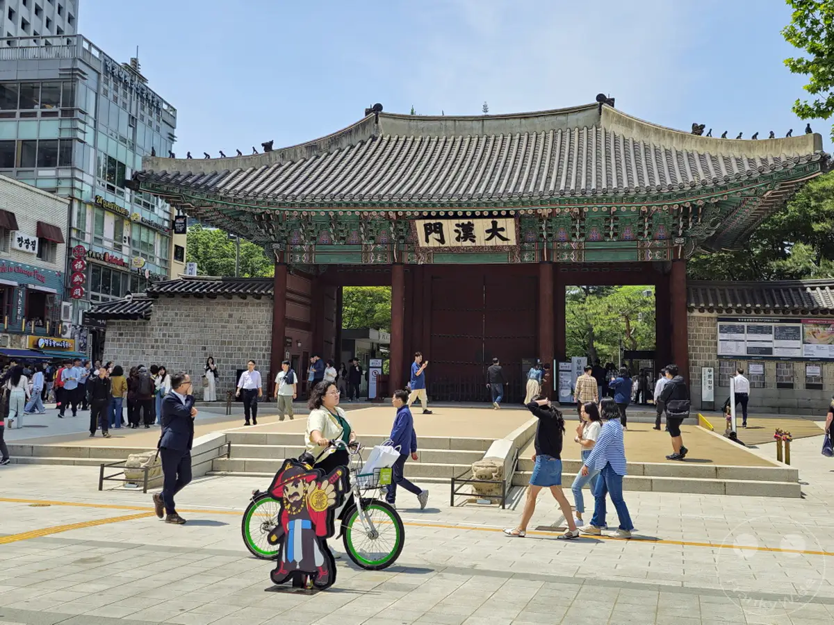 Ein belebter Platz vor dem Deoksugung Palace in Seoul, Südkorea, zeigt traditionelle und moderne Architektur. Touristen und Einheimische bewegen sich auf dem sonnigen Platz vor dem beeindruckenden Palasttor. Die Szene vermittelt eine lebhafte Atmosphäre des städtischen Lebens in Kombination mit historischem Erbe.