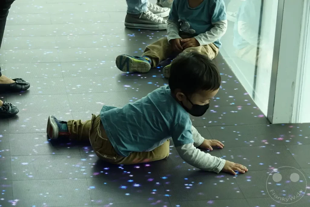 In Taipeh, Taiwan, erkunden Kinder eine interaktive Bodeninstallation vor dem berühmten Taipei 101. Bunte Lichter tanzen auf dem Boden und fesseln die Aufmerksamkeit der jungen Entdecker, die spielerisch und neugierig auf Entdeckungsreise gehen. Die Szene strahlt eine fröhliche und faszinierende Atmosphäre aus, die von der kindlichen Freude an der digitalen Kunst geprägt ist.