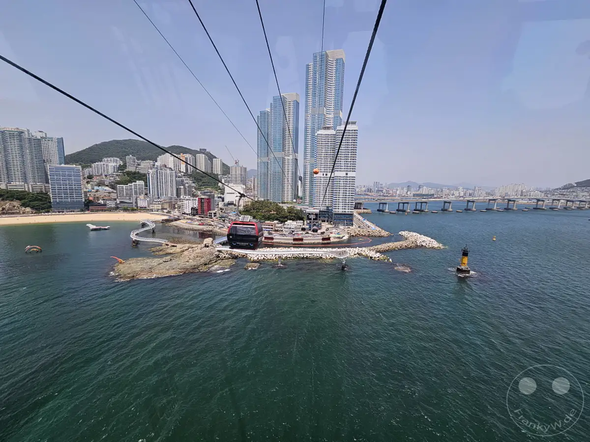 Die Seilbahn von Busan, Südkorea, schwebt über dem glitzernden Wasser mit Blick auf eine beeindruckende Skyline und eine weitläufige Brücke. Eine kleine Insel mit einem markanten roten Gondel und einem Leuchtturm bietet einen malerischen Kontrast zur modernen Stadtarchitektur. Die Szene strahlt eine Atmosphäre von Urlaub und Entspannung vor einer lebhaften urbanen Kulisse aus.