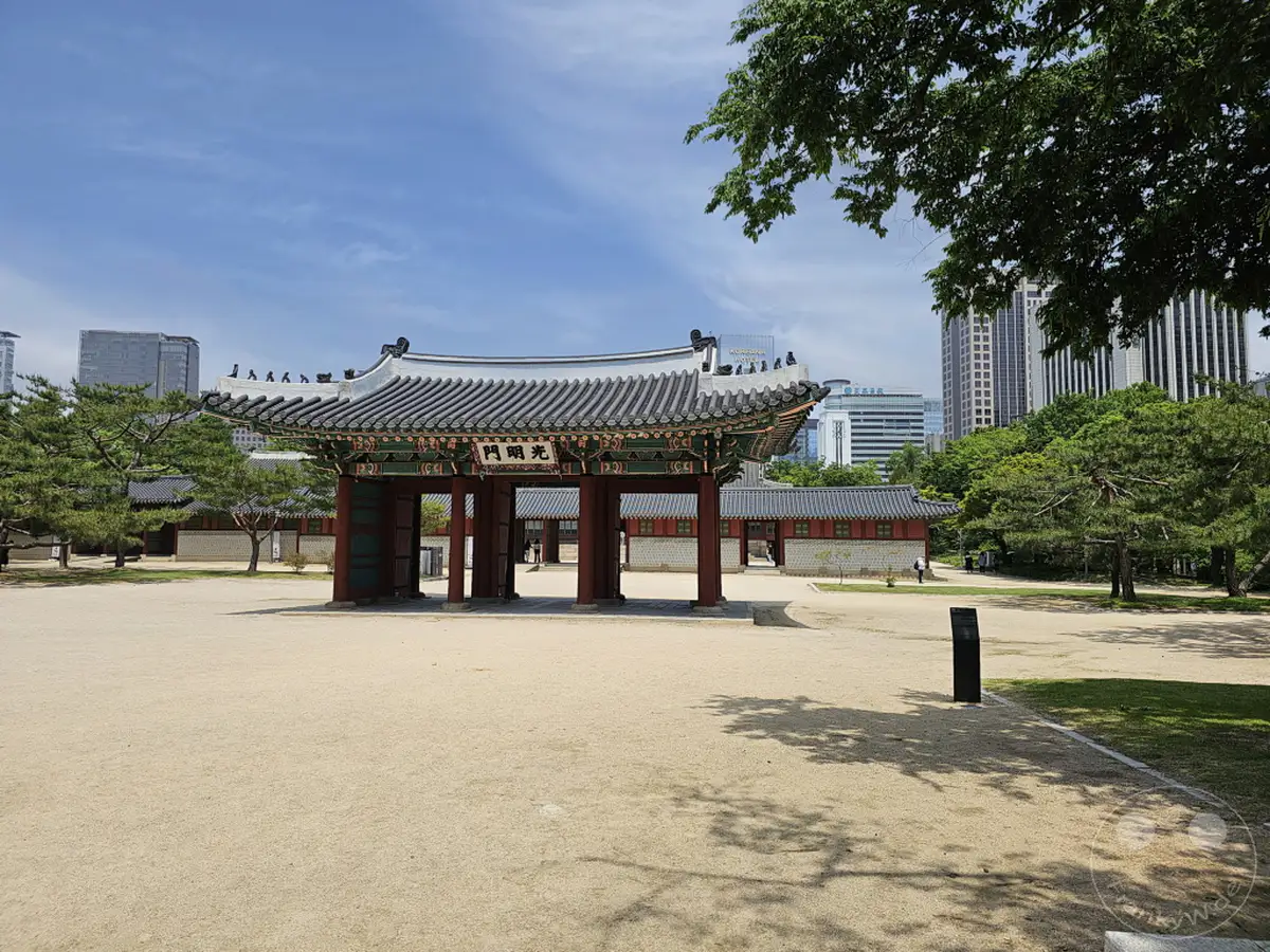 Ein traditionelles Tor steht im Vordergrund des Deoksugung Palace in Seoul, Südkorea, mit modernen Wolkenkratzern im Hintergrund. Helle Sonneneinstrahlung beleuchtet den sandigen Vorplatz und die üppig grünen Bäume, was eine harmonische Mischung aus Geschichte und Gegenwart schafft. Der weite, sonnige Himmel deutet auf einen klaren Sommertag in der pulsierenden Hauptstadt hin.