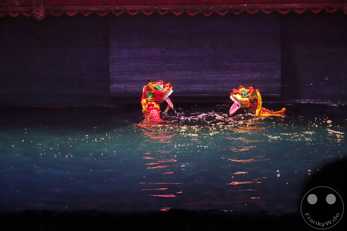 Zwei bunte Wasserpuppen in Drachenform bewegen sich auf einer dunklen Wasserbühne während einer traditionellen Aufführung von Wasserpuppen in Hanoi, Vietnam. Die Szene ist in gedämpftes Licht getaucht, das auf dem Wasser reflektiert und eine magische Atmosphäre schafft. Die kunstvollen Puppen sind aufwendig gestaltet und lassen auf eine aufwendige Inszenierung schließen.