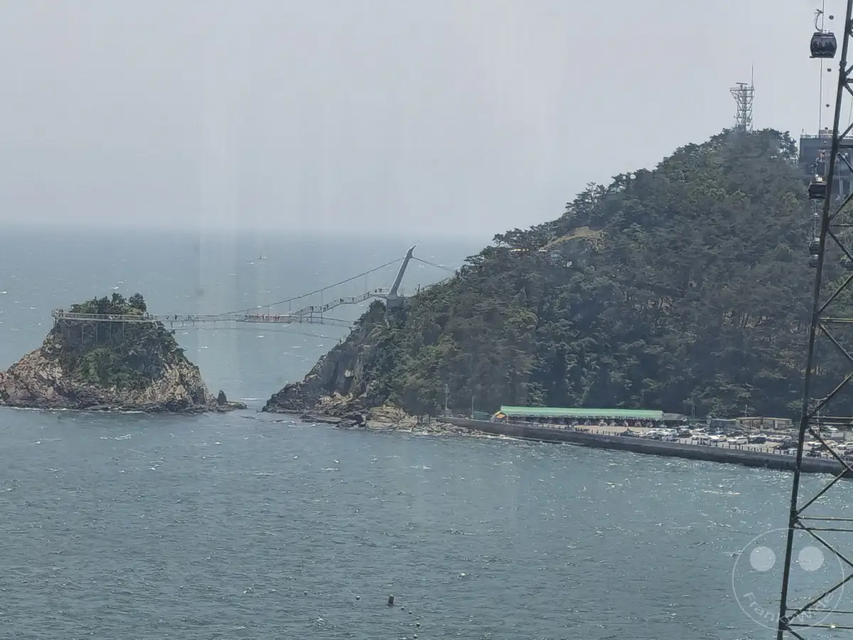 Die Seilbahn Songdo Cable Car in Busan, Südkorea, überspannt das Meer zwischen zwei felsigen Inseln. Eine beeindruckende Fußgängerbrücke verbindet die Inseln und bietet einen spektakulären Ausblick auf die Küste und das weite Meer. Die Szene strahlt eine friedliche Urlaubsatmosphäre aus, eingefangen an einem sonnigen Tag.