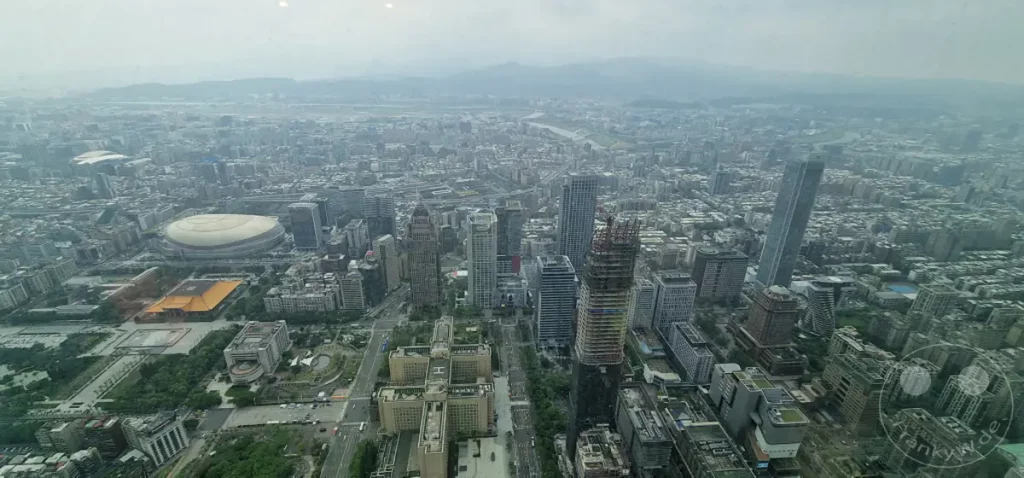 Ein weitläufiger Blick über die Metropole Taipeh in Taiwan, dominiert von modernen Hochhäusern und dem ikonischen Taipei 101. Die Stadt erstreckt sich bis zu den nebligen Bergen am Horizont, was eine leicht gedämpfte, urbane Atmosphäre erzeugt. Auf dem Boden sind einige öffentliche Gebäude und Grünflächen erkennbar, die das geschäftige Stadtleben ergänzen.