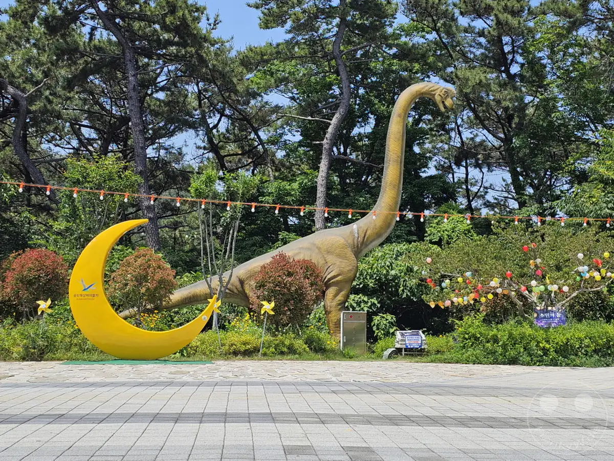 Im Songdo Sky Park in Busan, Südkorea, steht eine beeindruckende Dinosaurier-Skulptur neben einer großen, gelben Mondsichel-Dekoration. Bunte Lichterketten und kleine Windräder schmücken die Anlage und schaffen eine verspielte Atmosphäre inmitten der grünen Natur. Das sonnige Wetter und die detailreichen Skulpturen laden zum Erkunden und Fotografieren ein.