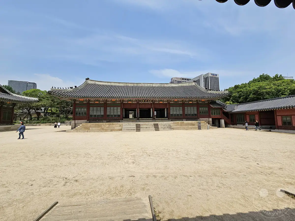 Ein weitläufiger Sandplatz öffnet sich vor dem Hauptgebäude des Deoksugung Palastes in Seoul, Südkorea. Die traditionelle koreanische Architektur mit ihren geschwungenen Dächern und roten Holzelementen bildet einen starken Kontrast zum blauen Sommerhimmel. Einige Besucher spazieren auf dem Hof, während moderne Gebäude und grüne Bäume im Hintergrund zu sehen sind.