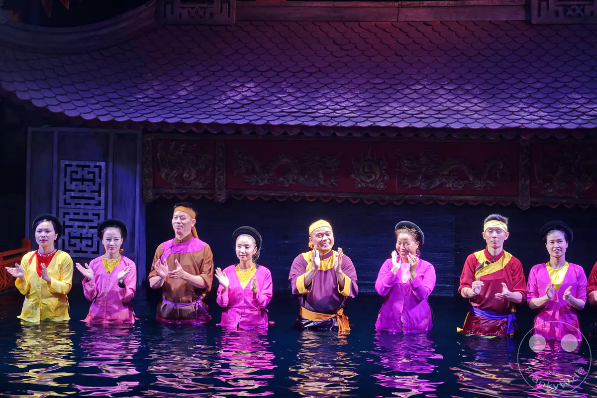 In Hanoi, Vietnam, wird die traditionelle Kunstform des Wasserpuppenspiels aufgeführt, bei der Darsteller in bunten Kostümen bis zur Hüfte in Wasser stehen. Sie führen eine Aufführung mit geklatschten Händen vor einer Kulisse mit Drachenmotiven und traditioneller Architektur auf. Die spiegelnden Reflexionen im Wasser erzeugen eine magische und lebendige Atmosphäre, die die kulturelle Darbietung unterstreicht.