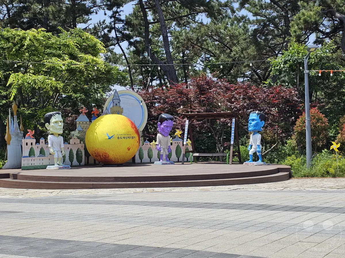 Auf einer Holzplattform im Songdo Sky Park in Busan, Südkorea, stehen drei auffällige Skulpturen, die an Frankenstein erinnern. Eine große, gelb-rote Kugel mit koreanischer Schrift dominiert die Mitte, flankiert von lila und blauer Frankenstein-Figur, während im Hintergrund üppiges Grün und Bäume zu sehen sind. Die Szene wirkt verspielt und einladend und deutet auf einen unterhaltsamen öffentlichen Raum hin.