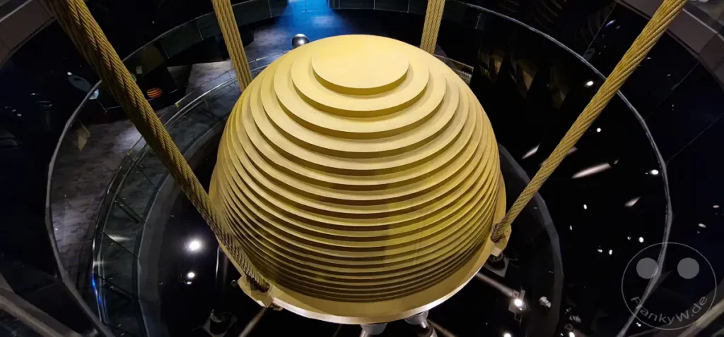 Der riesige, goldene Massenausgleichsdämpfer aus konzentrischen Ringen hängt in der Mitte des Taipei 101 Wolkenkratzers in Taipeh, Taiwan. Starke Stahlseile halten die massive Kugel, während die umliegenden Ebenen des Gebäudes im dunklen, reflektierenden Inneren sichtbar sind. Die Szene vermittelt ein Gefühl von Ingenieurskunst und visueller Dramatik im Herzen eines ikonischen Wahrzeichens.